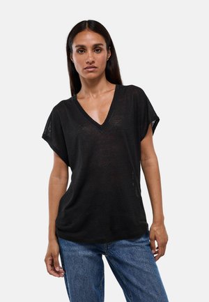 Femme aux longs cheveux foncés, portant un haut noir ample à manches courtes et col en V, et un jean bleu, debout devant un fond uni.