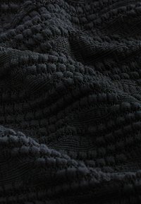 Tissu noir texturé avec un motif tissé, présentant des points en relief et des rayures lisses dans des tons sombres, créant une apparence douce et dense.