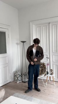 Brown, fuzzy jacket with two black zippered pockets, worn over a white tank top, paired with blue jeans and gray shoes. Simple interior backdrop.
