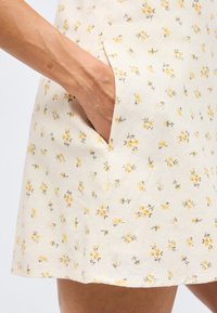 Χέρι στην τσέπη φούστας σε κρεμ χρώμα με μικρό κίτρινο λουλουδένιο μοτίβο, εμφανίζοντας τις φλέβες του βραχίονα και την υφή του δέρματος.