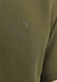 Olivgrön texturerad polo med rutmönster, prydd med en broderad sköldlogotyp på bröstet och korta ärmar.