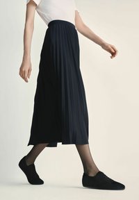 Czarna plisowana spódnica z wysokim pasem, noszona z czarnymi butami. Tkanina ma gładką strukturę i sięga do połowy łydki.