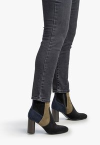 Mujer que lleva botas de ante negras, marrones y azul marino con tacones en bloque, combinadas con jeans ajustados gris oscuro, sobre un fondo blanco.