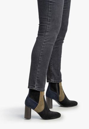 Femme portant des bottines en suède noires, marron et bleu marine à talons carrés, associées à un jean skinny gris foncé sur fond blanc.
