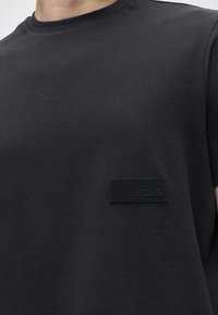 Κοντινό πλάνο προσώπου που φοράει μαύρο t-shirt με στρογγυλή λαιμόκοψη και μαύρο ορθογώνιο μπαλώμα με την ένδειξη "BOSS" στο στήθος.