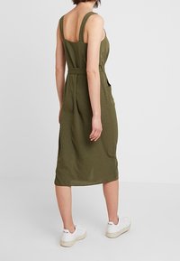 Abito senza maniche verde oliva con vita aderente, scollatura sulla schiena e tasche laterali; abbinato a sneaker sportive bianche.