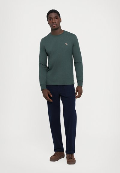 Man som bär en mörkgrön långärmad tröja med liten logotyp, marinblåa byxor och bruna sneakers, stående mot en vit bakgrund.