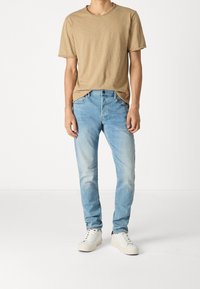 Person som bär en beige kortärmad T-shirt, ljusblå jeans och vita sneakers som står mot en neutral vit bakgrund.