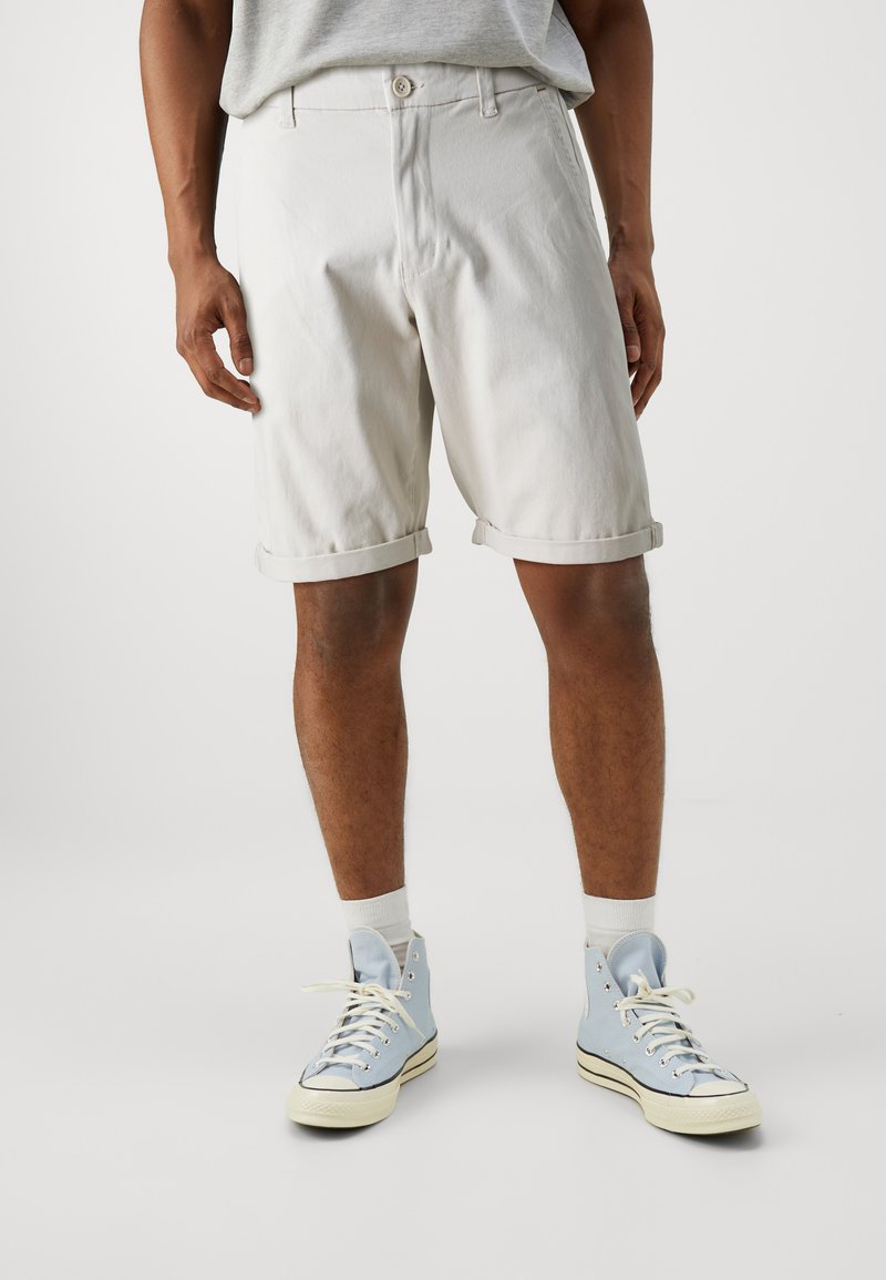 Pantalones cortos de mezclilla blanca con puños enrollados, que cuentan con una bragueta de botones y bolsillos laterales, combinados con zapatillas altas azul claro y calcetines blancos.