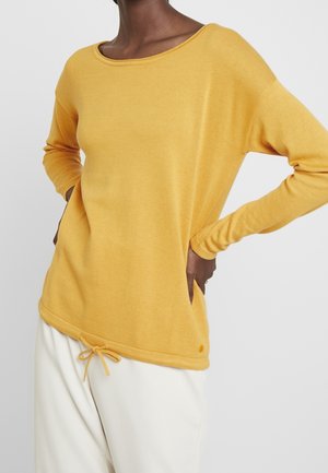 Personne portant un haut à manches longues jaune moutarde avec un cordon à l'ourlet et un pantalon blanc, mains posées sur les hanches sur un fond uni.