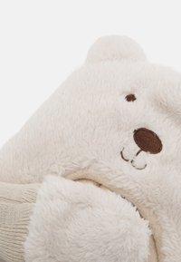 Weiße plüschige Hausschuhe in Form eines Bären mit weicher Textur, ausgestattet mit einem braunen, gestickten Gesicht und einem beigen Bündchen am Einstieg.