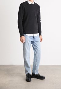 Schwarzer Baumwollpullover über einem weißen Kragenhemd, kombiniert mit hellblauen Jeans und schwarzen Lederschuhen mit dicker Sohle.