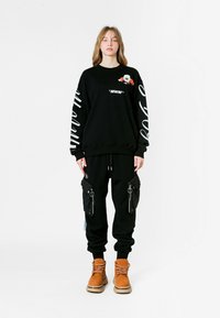 Sudadera negra con detalles gráficos y de texto, combinada con pantalones cargo negros con bolsillos con cremallera, usados con botas de cordones color beige.