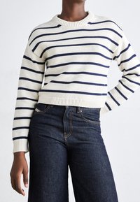 Pull à manches longues crème avec des rayures horizontales bleu marine, col et poignets côtelés, associé à un jean en denim bleu foncé.