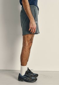 Pantalones cortos de felpa grises con cordón ajustable, bolsillos laterales y un parche de logo. Llevados con zapatillas deportivas negras y azules y calcetines blancos.