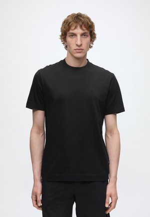 Jeune homme aux cheveux bouclés portant un t-shirt noir uni à manches courtes et un pantalon noir, debout devant un fond blanc uni.