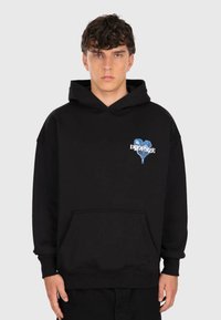 Young man with curly hair wearing a black hoodie with a blue heart and white "DROPSIZE" text on the chest, standing against a plain background.