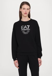 Fekete pulóver EA7 Emporio Armani fehér logóval. Jellemzője a kerek nyakú szabás, hosszú ujjak, valamint az elasztikus mandzsetták és alsó szegély.