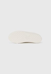 Hvit sneakersåle med teksturert gripe mønster og preget "CK"-logo plassert i sentrum på en enkel lys bakgrunn.