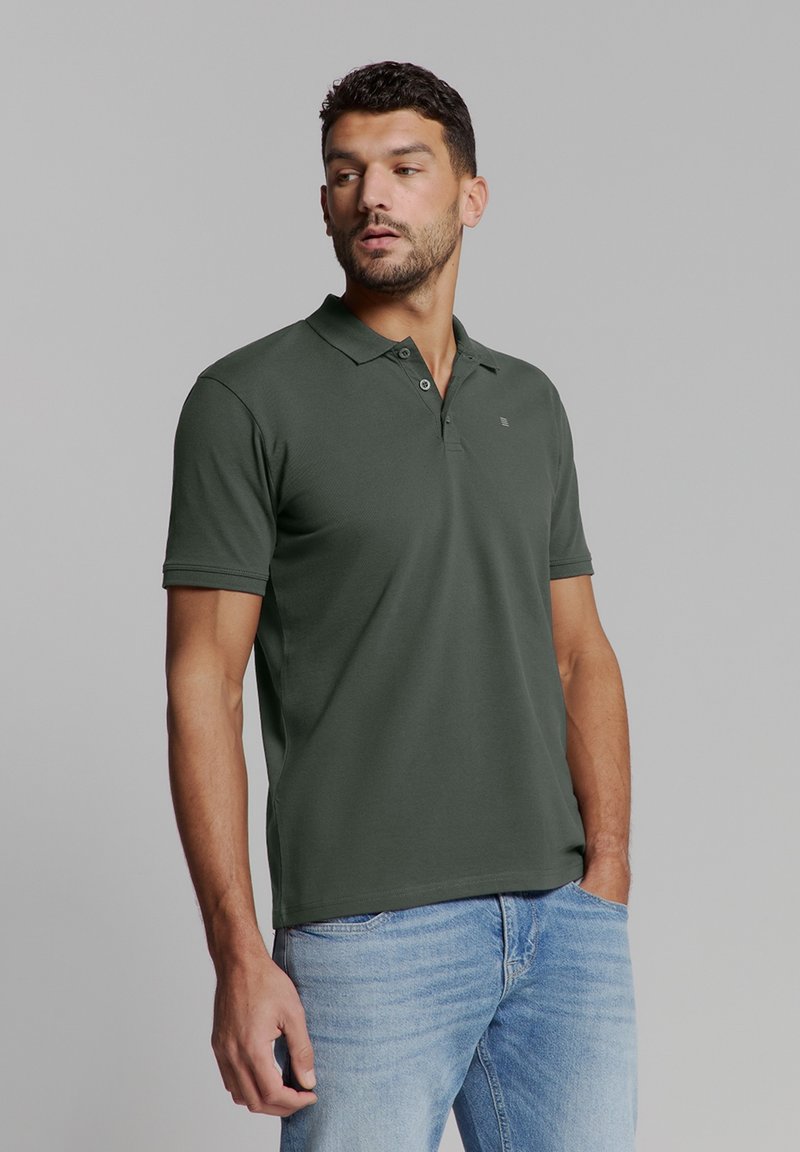 Donkergroene polo met kraag en twee knopen, korte mouwen, gladde textuur, aangevuld met lichtblauwe denim jeans.