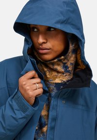 Blauwe waterdichte jas met een capuchon, met een ritssluiting en klittenbandsluitingen bij de manchetten. Onder de jas is een gestreepte, wollen sjaal zichtbaar.