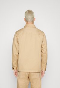 Beige bavlněná bunda s límečkem, manžetami na knoflík a zadním sedlem. Tkanina má hladkou texturu a volnější střih, nošena s odpovídajícími kalhotami.