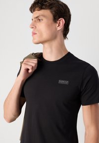 Tricou din bumbac negru cu mâneci scurte, având un mic logo Barbour International pe piept. Modelul ține o cămașă pliata de culoare verde măslinie.