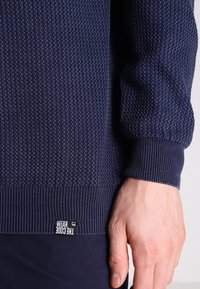 Pull en maille bleu marine avec un motif texturé, poignets et ourlet côtelés. Présente une petite étiquette indiquant "WEAR GOOD" en bas.
