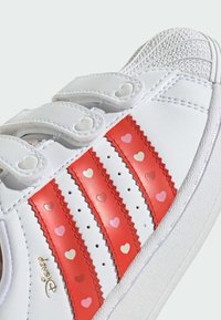 Biele tenisky so pruhmi s červeným srdcom. Majú lemovanie s vlnitým okrajom, perforácie pre priedušnosť a suché zipsy. Textúrovaná gumová podrážka.