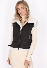 Zwarte leren vest met een witte nepbont kraag, ritsluiting, verstelbare banden en zakken. Het model draagt een crèmekleurige coltrui jurk.