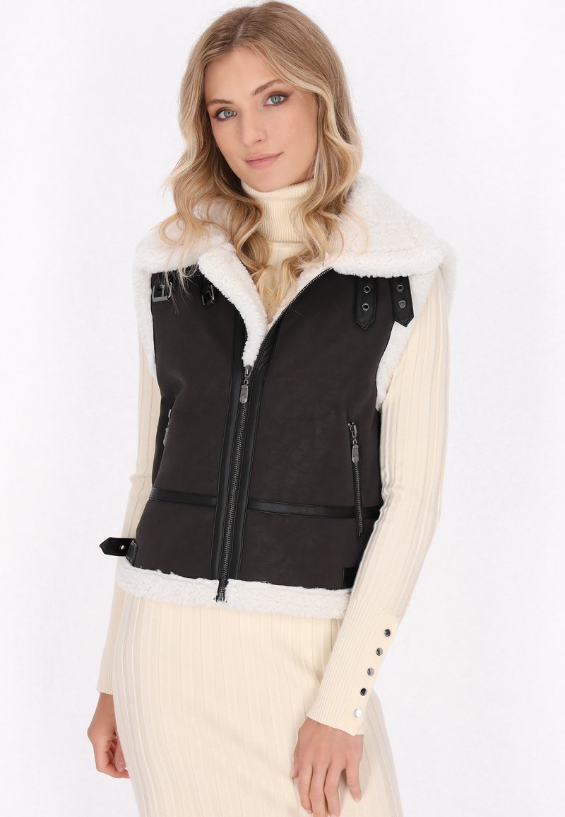 Zwarte leren vest met een witte nepbont kraag, ritsluiting, verstelbare banden en zakken. Het model draagt een crèmekleurige coltrui jurk.