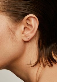 Pendientes de oro que presentan un pequeño pendiente y una cadena larga y delgada con un acento rectangular, colgando elegantemente cerca de la oreja.