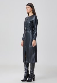 Robe en cuir noir au design cintré, avec des manches longues, une taille ceinturée et une fente à l'avant. Assortie à des bottes brillantes jusqu'aux genoux.