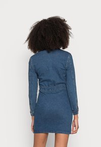 Jeanimekko tummansinisellä värillä, pitkähihaisilla, vyötäröltä muotoillulla ja hieman levenevällä helmalla. Varusteena kaulus ja napitettavat hihat.