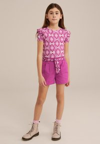 Roza vzorčasta majica s kratkimi rokavi in volančki, v kombinaciji z visokimi roza hlačami. Tan čevlji s čipkastimi vezalkami in črtaste nogavice dopolnjujejo videz.