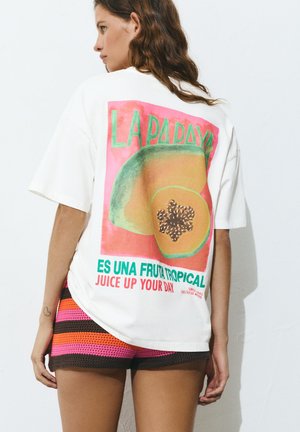 Femme portant un T-shirt blanc oversize avec un imprimé fruit de papaye et un texte en espagnol et en anglais, associé à un short tricoté rayé coloré.