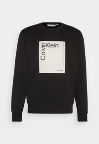 Felpa nera con maniche lunghe, collo rotondo e una grafica rettangolare bianca con "Calvin Klein" e "Est. 1968" in grassetto nero.