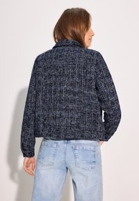 Giacca texture navy con design cropped e maniche lunghe, caratterizzata da una leggera lucentezza e cuciture a vista in contrasto, abbinata a jeans in denim chiaro.