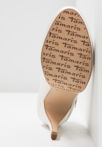 Chaussure blanche à talon haut, inclinée pour montrer la semelle avec le logo "Tamaris" répété en marron sur un fond beige clair.