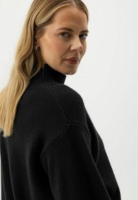 Blonde Frau mit langen Haaren, die einen schwarzen Rollkragenpullover trägt und über ihre Schulter auf einen schlichten hellen Hintergrund schaut.