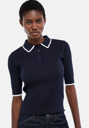 Femme noire portant un polo côtelé bleu marine ajusté avec un bord blanc sur le col et les manches, associé à un jean bleu foncé, regardant devant elle.