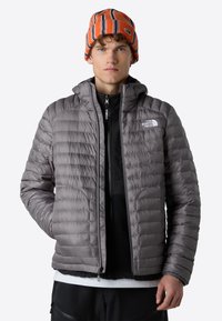 Grå puffjacka med huva, med quiltad textur och en logotyp på vänstra bröstet. Bärs över en svart zip-up tröja och matchas med en randig mössa.