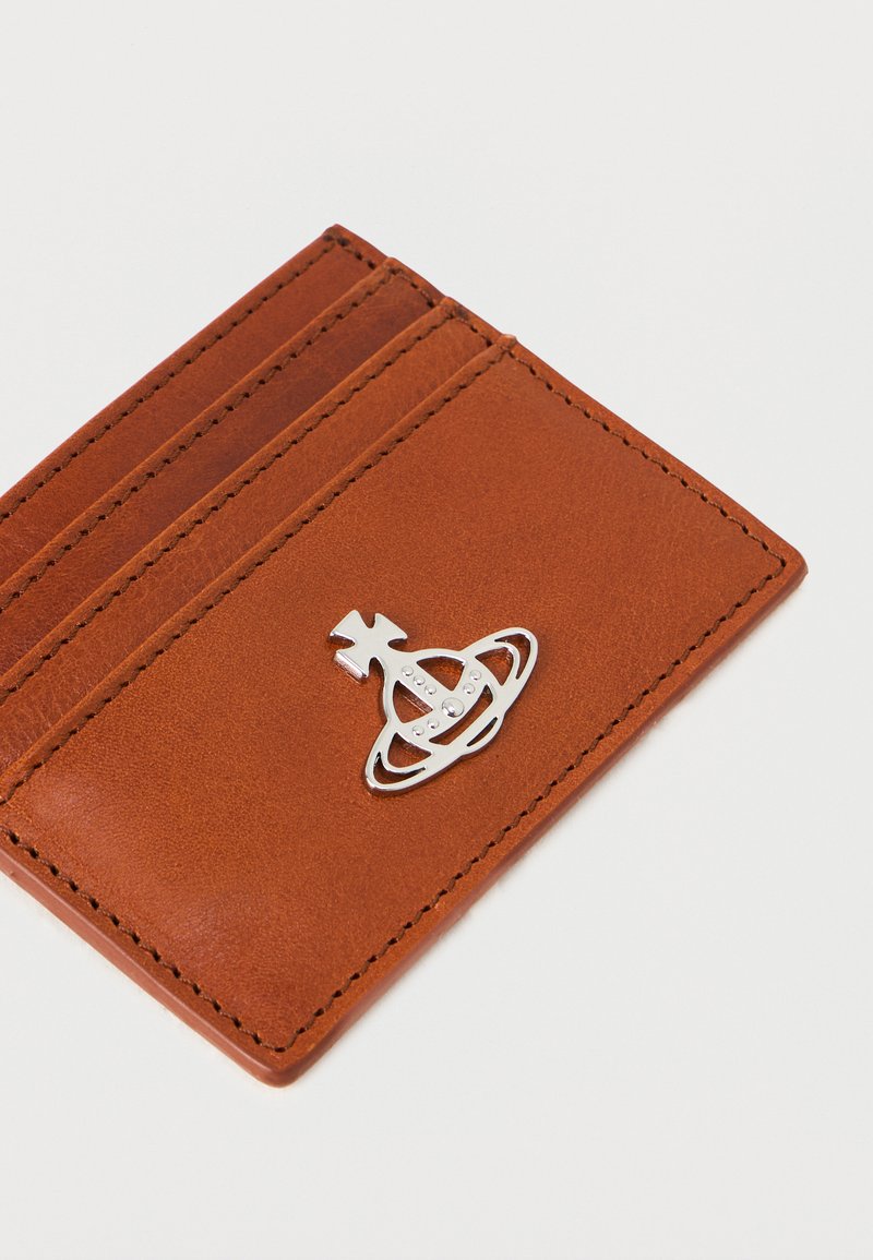 Tarjetero de cuero marrón con tres ranuras y un acento de logo plateado con un diseño de planeta. Textura suave con costuras en contraste.