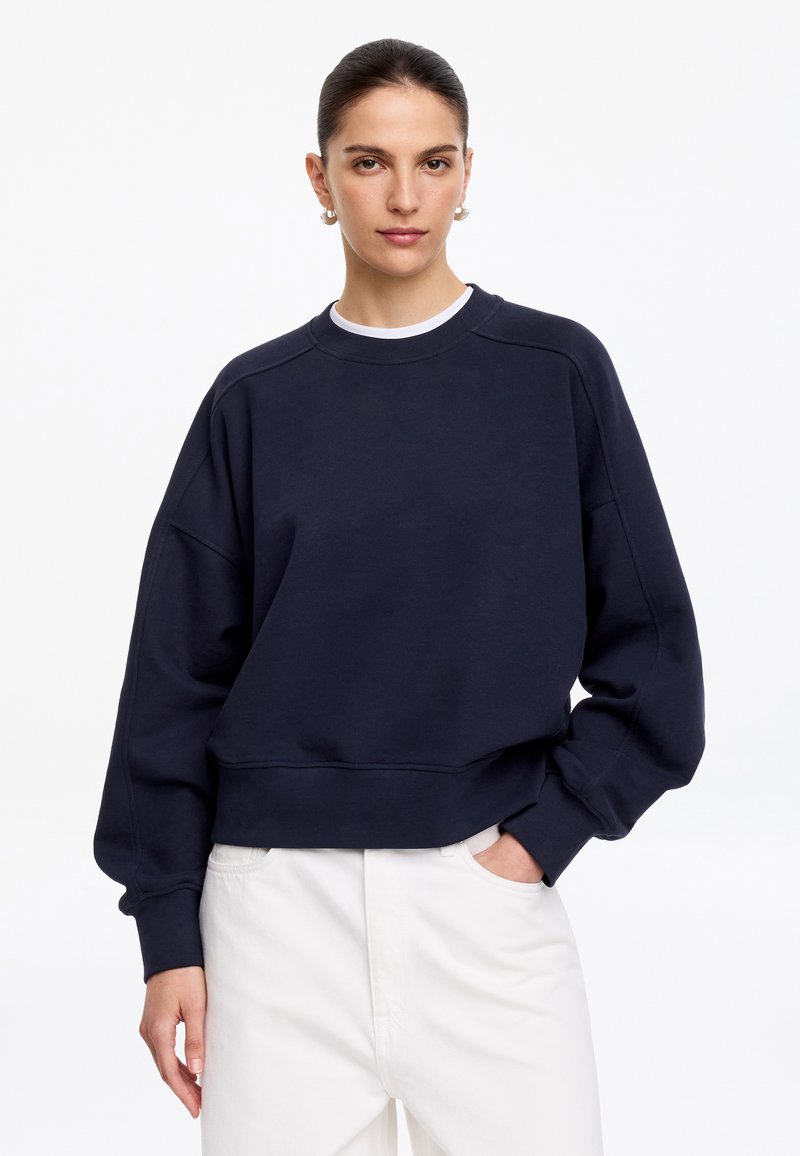 Femme aux cheveux foncés attachés en queue de cheval, portant des boucles d’oreilles créoles en or, un sweat-shirt bleu marine et un pantalon blanc, debout avec une main dans la poche.