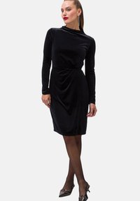 Czarna aksamitna sukienka z długimi rękawami, okrągłym dekoltem i drapowanym pasem. Charakteryzuje się dopasowanym krojem i długością do kolan.
