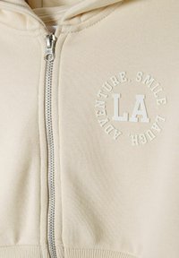 Sudadera beige con cremallera, hecha de una suave mezcla de algodón, con un diseño gráfico redondo que incluye "LA" y texto. Incluye cremallera plateada y puños acanalados.