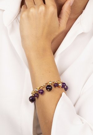 Bracciale dorato con una miscela di perline viola e nere, accentuato da orbite metalliche, indossato su un polso contro uno sfondo bianco.