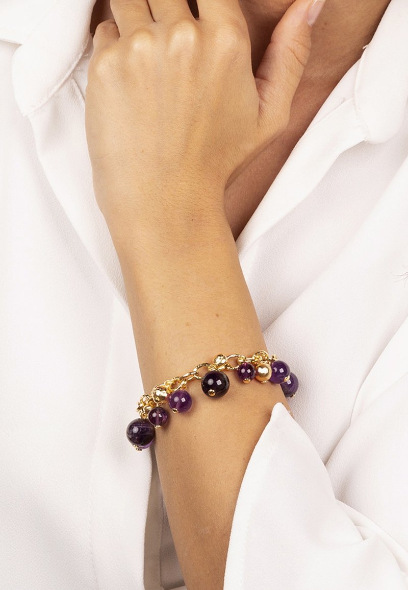 Bracciale dorato con una miscela di perline viola e nere, accentuato da orbite metalliche, indossato su un polso contro uno sfondo bianco.