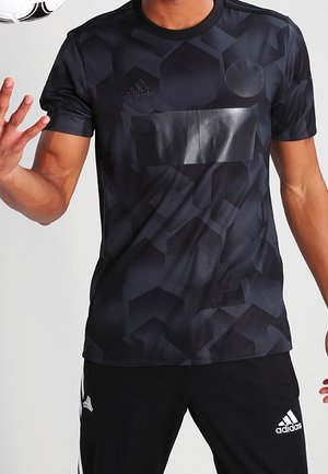 Homme portant une tenue de sport Adidas noire avec des motifs géométriques sur le maillot et tenant un ballon de football noir et blanc près de son épaule.