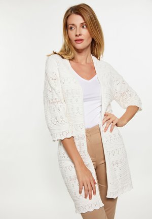 White open-front cardigan with intricate lace detailing, short sleeves, and scalloped edges, worn over a white top and beige pants.